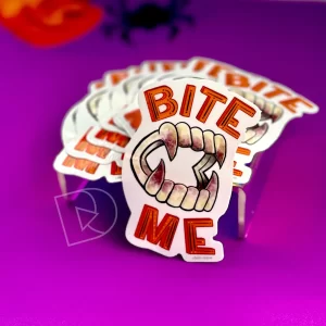Sticker: Illustrated pair of bloodied false vampire teeth surrounded by the phrase "Bite Me" in orange hand-lettering, photographed on a purple background.